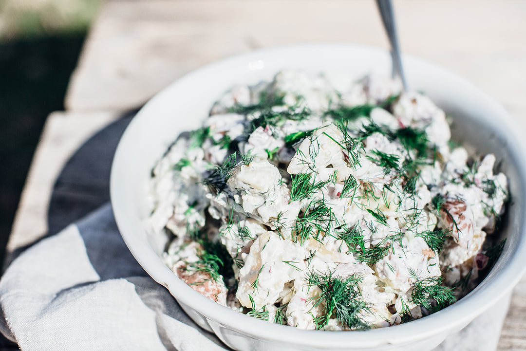 A creamy potato salad topped with fresh dill in a white serving bowl on an outdoor picnic table