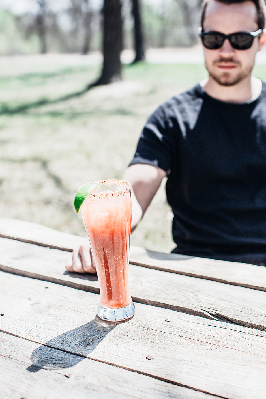 A man reaching for a caesar on a picnic table. He is wearing a black t-shirt and sunglasses.