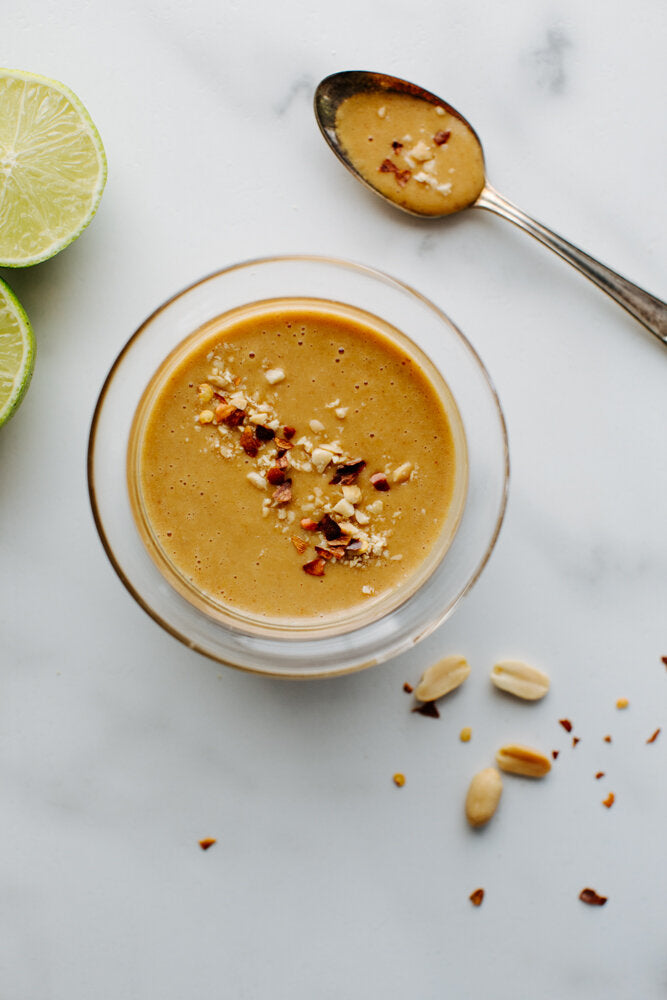 A glass bowl filled with Tangy Peanut Dressing on a marble table with a silver spoon and lime beside it