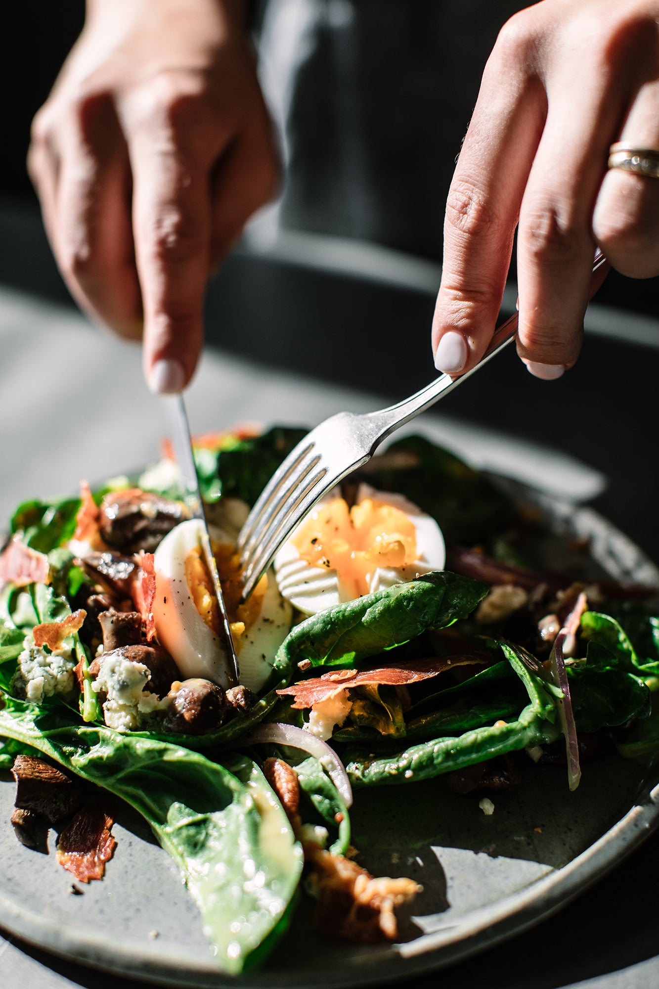 Honey Mustard Vinaigrette Dressed Spinach Salad w/ Crispy Prosciutto and hand holding knife and fork cutting into egg