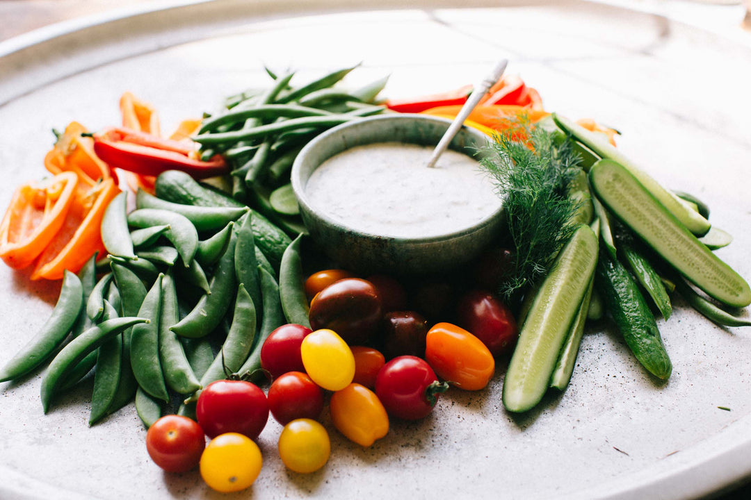 A bowl of Smak Dab's Dilly Ranch Dip surrounded by fresh sliced cumber, cherry tomatoes, snap peas, and cut bell peppers