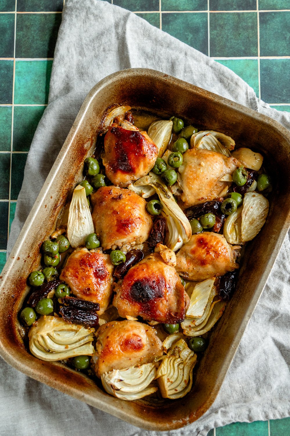 Mediterranean Chicken Marbella with Orange, Olives, Fennel and Dates on a green tile in a copper baking pan