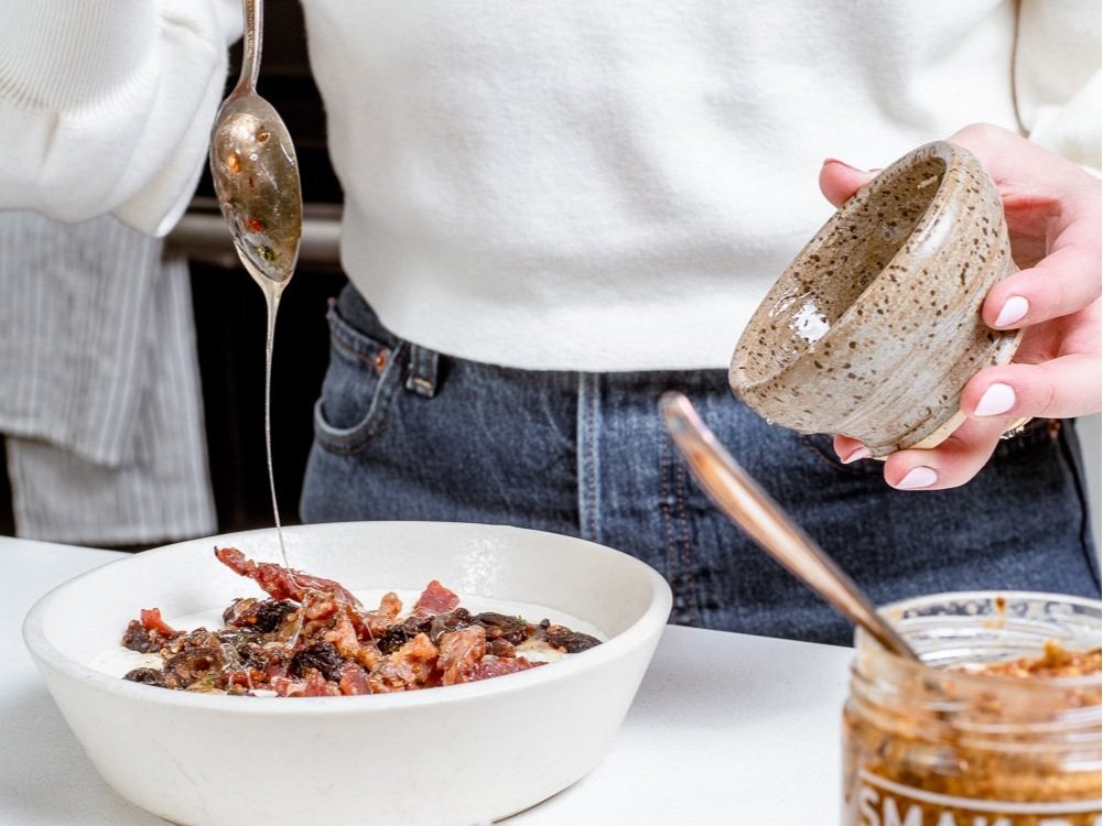 Whipped Goats Cheese Dip with candied dates and bacon, woman drizzling maple mustard and honey over dip in a white bowl