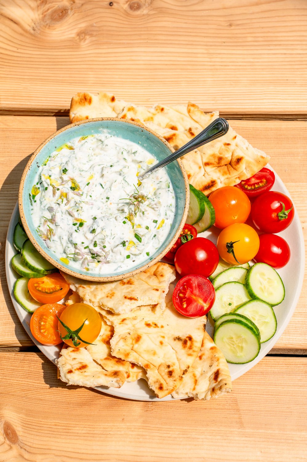 A bowl filled with Dill Pickle Feta dip on a plate with fresh tomatoes, pita bread, and sliced cucumber on a picnic table