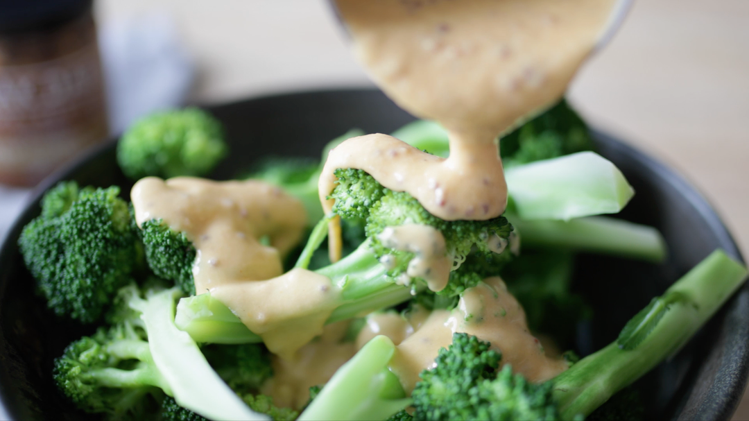 Broccoli being drizzled with Mustard Cheese Sauce in a black bowl