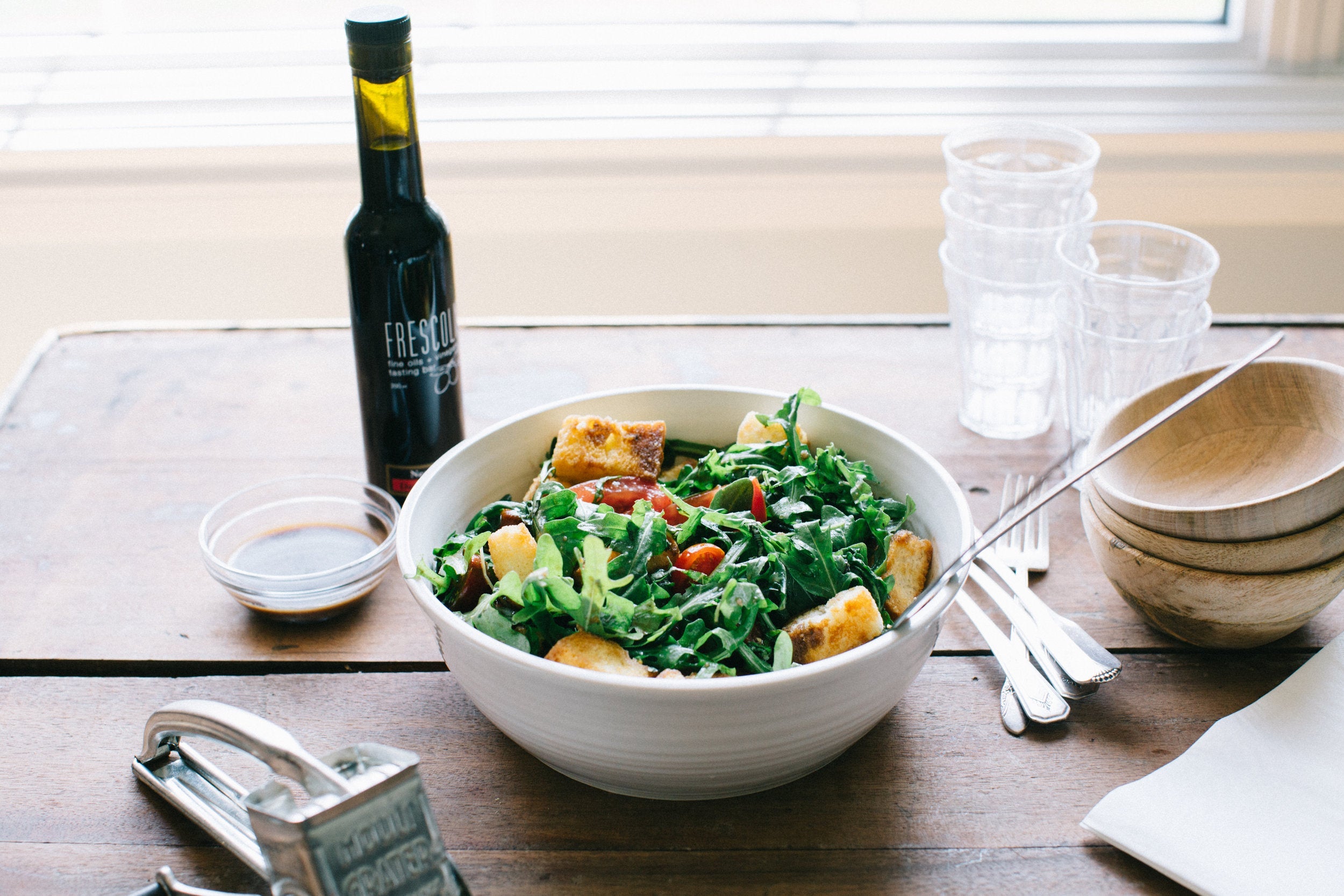 A Tomato Panzanella Salad with Balsamic Dijon Dressing in a white bowl on a wooden table with serving utensils in the bowl. Beside the bowl is 3 wooden bowls and 3 water glasses.